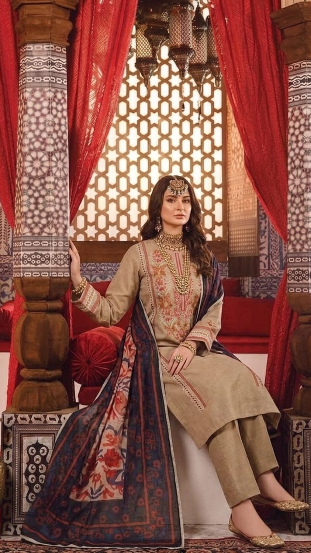 Woman in traditional attire sitting in a decorated room with red curtains and intricate patterns.
