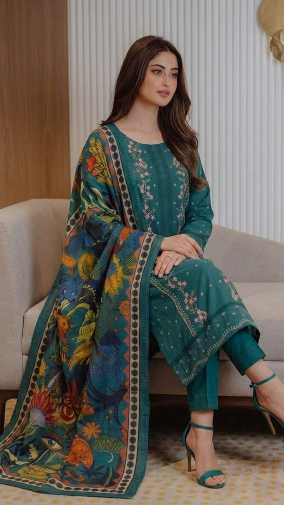 Woman wearing a teal embroidered outfit with a colorful dupatta, sitting on a couch.