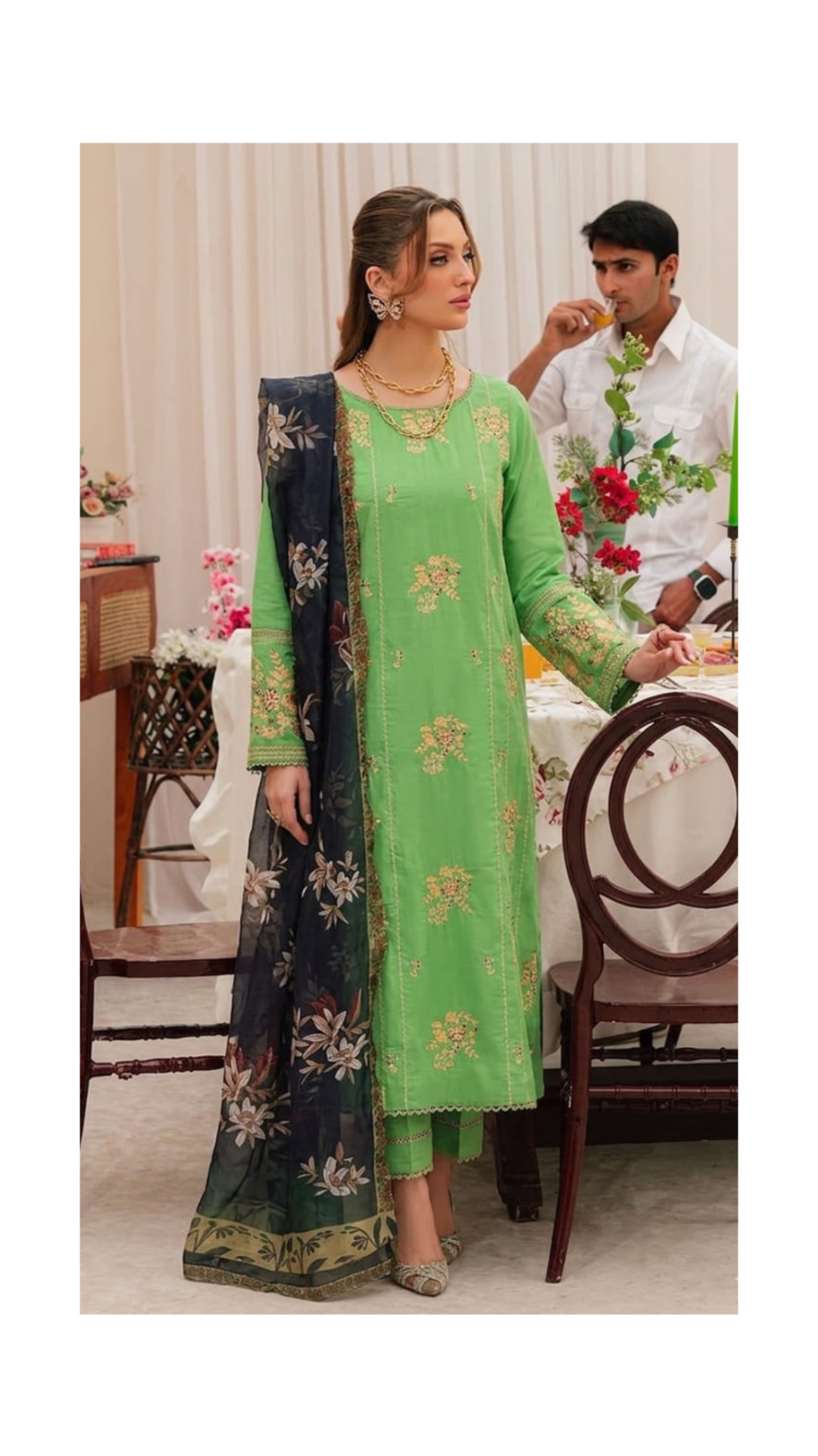 Woman in a green embroidered outfit with a man in the background at a formal event.