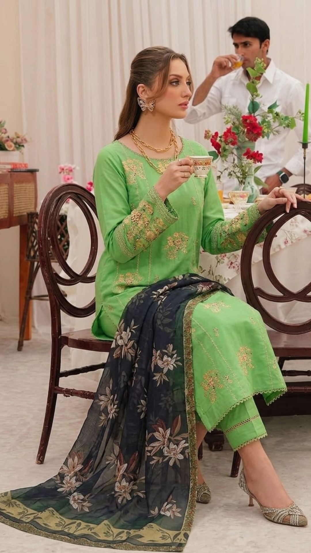 Woman in a green and navy traditional outfit sitting at a table with a man in the background.