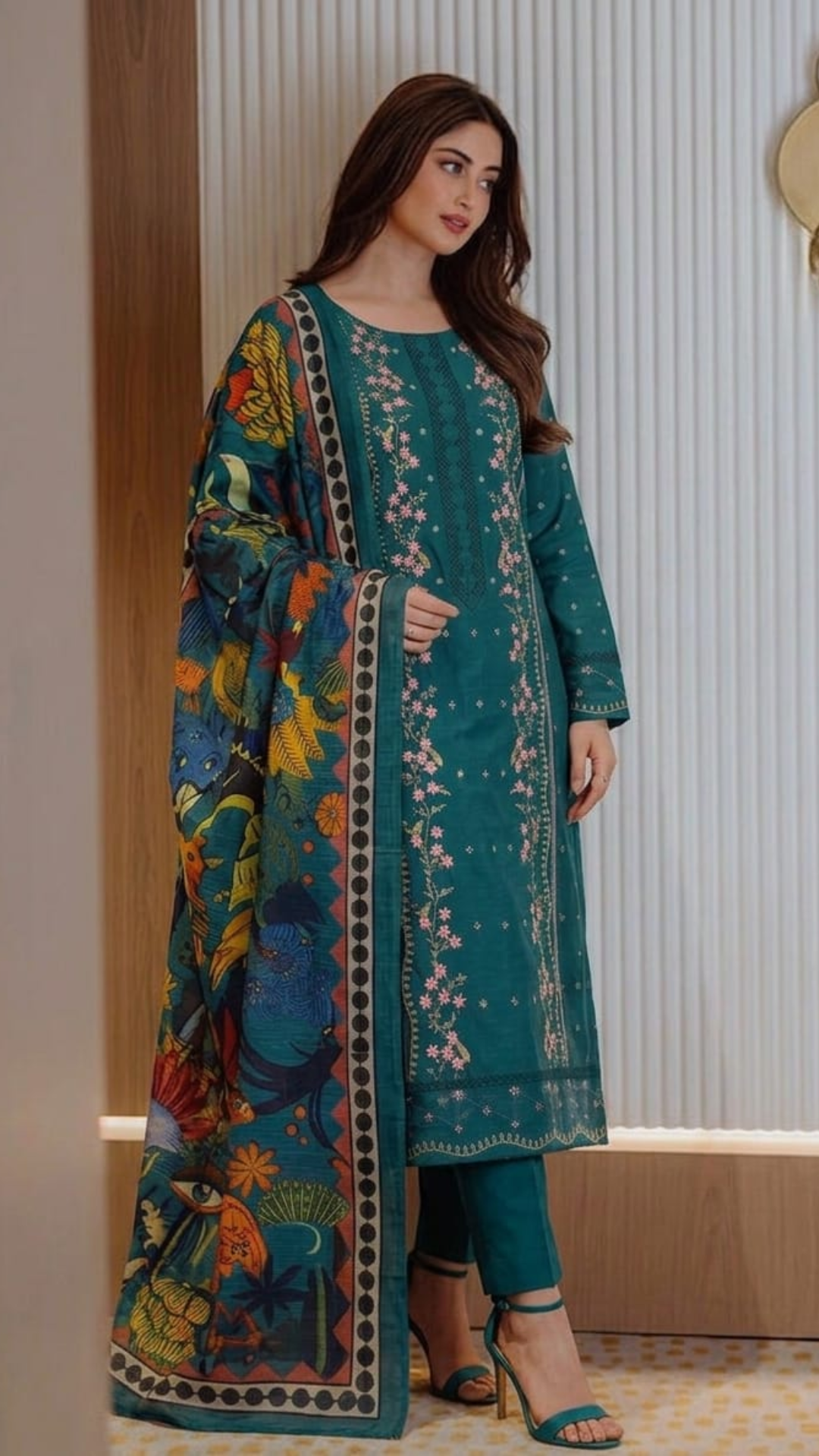 Woman wearing a teal embroidered outfit with a colorful jacket indoors.
