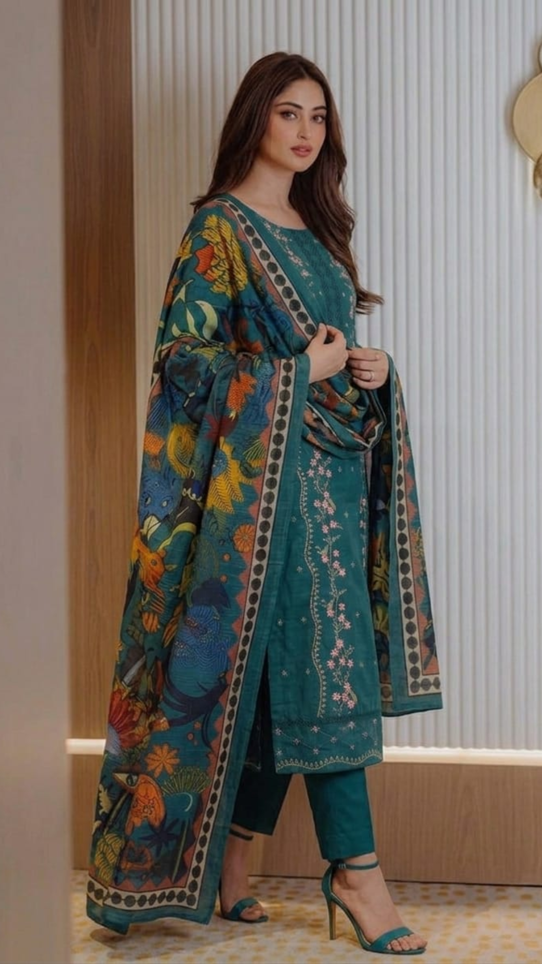 Woman wearing a teal embroidered outfit with a colorful pattern in an indoor setting.